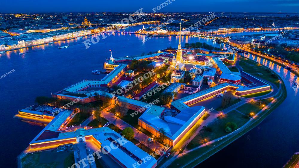 Панорамный ночной вид на Санкт-Петербург с высоты дрона с Петропавловской крепостью и рекой Невой в огнях вечернего города. Исторический центр северной столицы России, подсвеченные набережные, отражения в воде и масштаб городской архитектуры создают впечатляющий городской пейзаж. Аэрофотосъёмка подчёркивает красоту ночного Петербурга, атмосферу белых ночей и величие архитектурного наследия города. Изображение идеально подходит для публикаций о путешествиях по России, ночной архитектуре Санкт-Петербурга, туристических достопримечательностях и современных видах города с высоты.